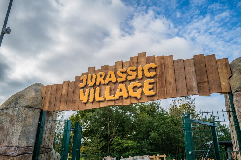 A wooden sign reading "Jurassic Village" set against a blue sky with trees below.
