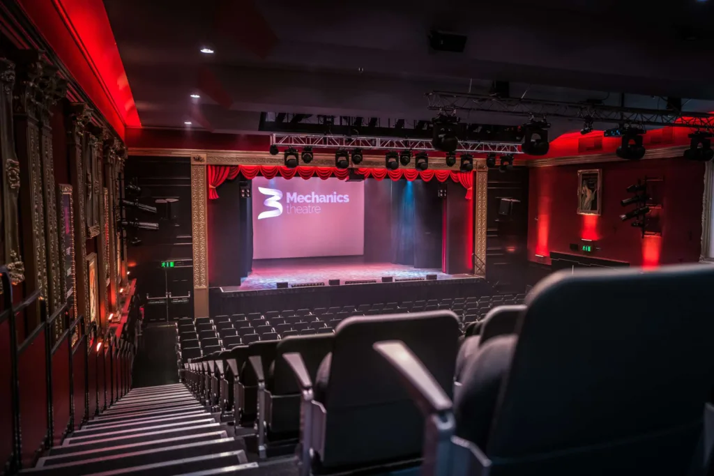 The interior of Mechanics Theatre, showing an ornate stage with red drapery, overhead lighting trusses, and empty auditorium seating.