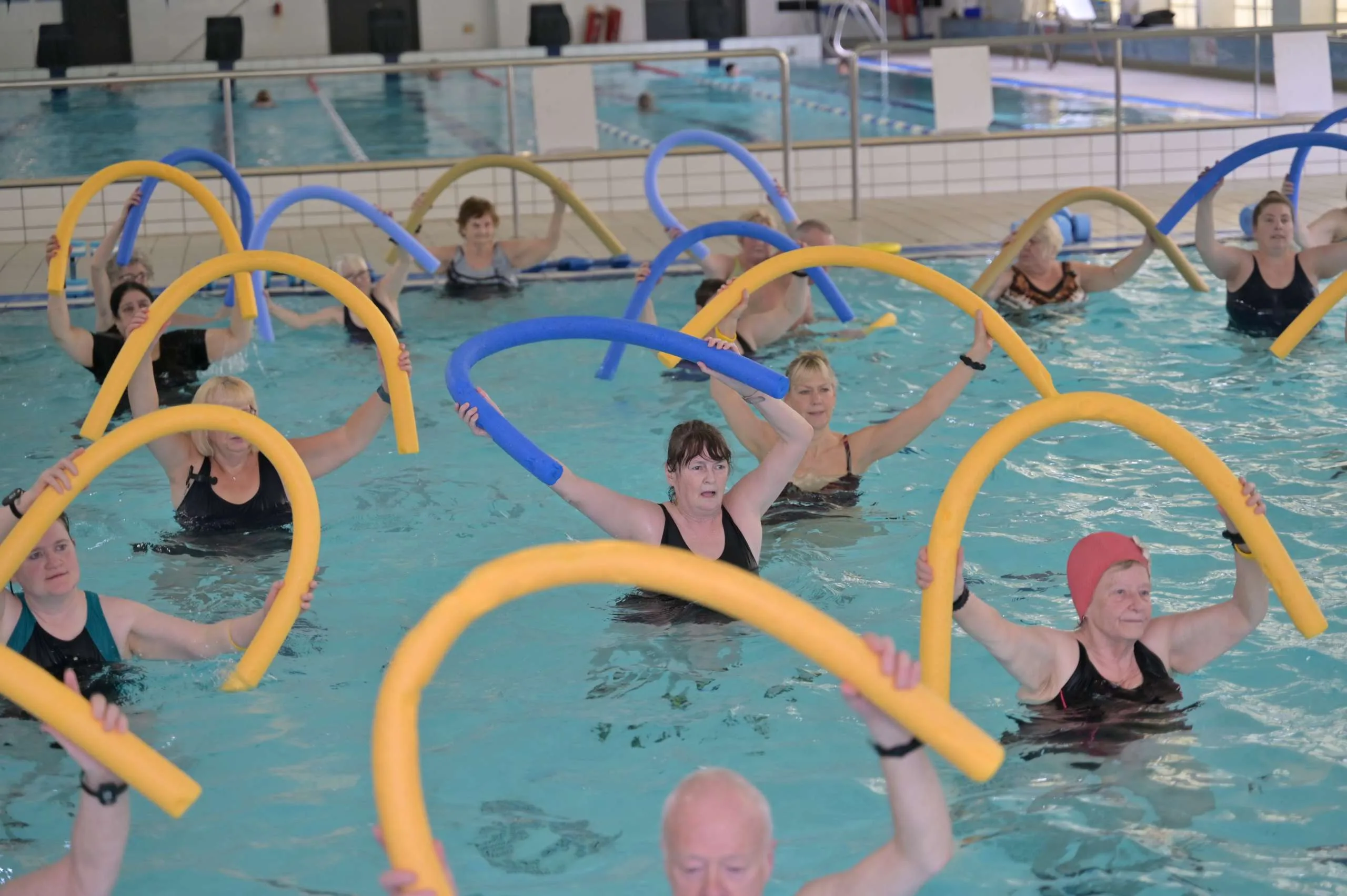 A group of adults taking part in an aqua aerobics class in an indoor swimming pool, holding foam noodles above their heads