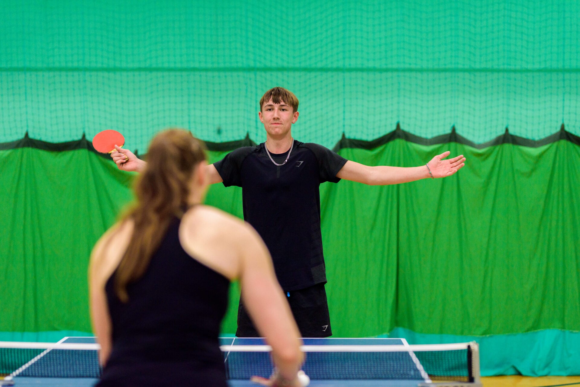 A young man and woman play table tennis on an indoor court with a green backdrop