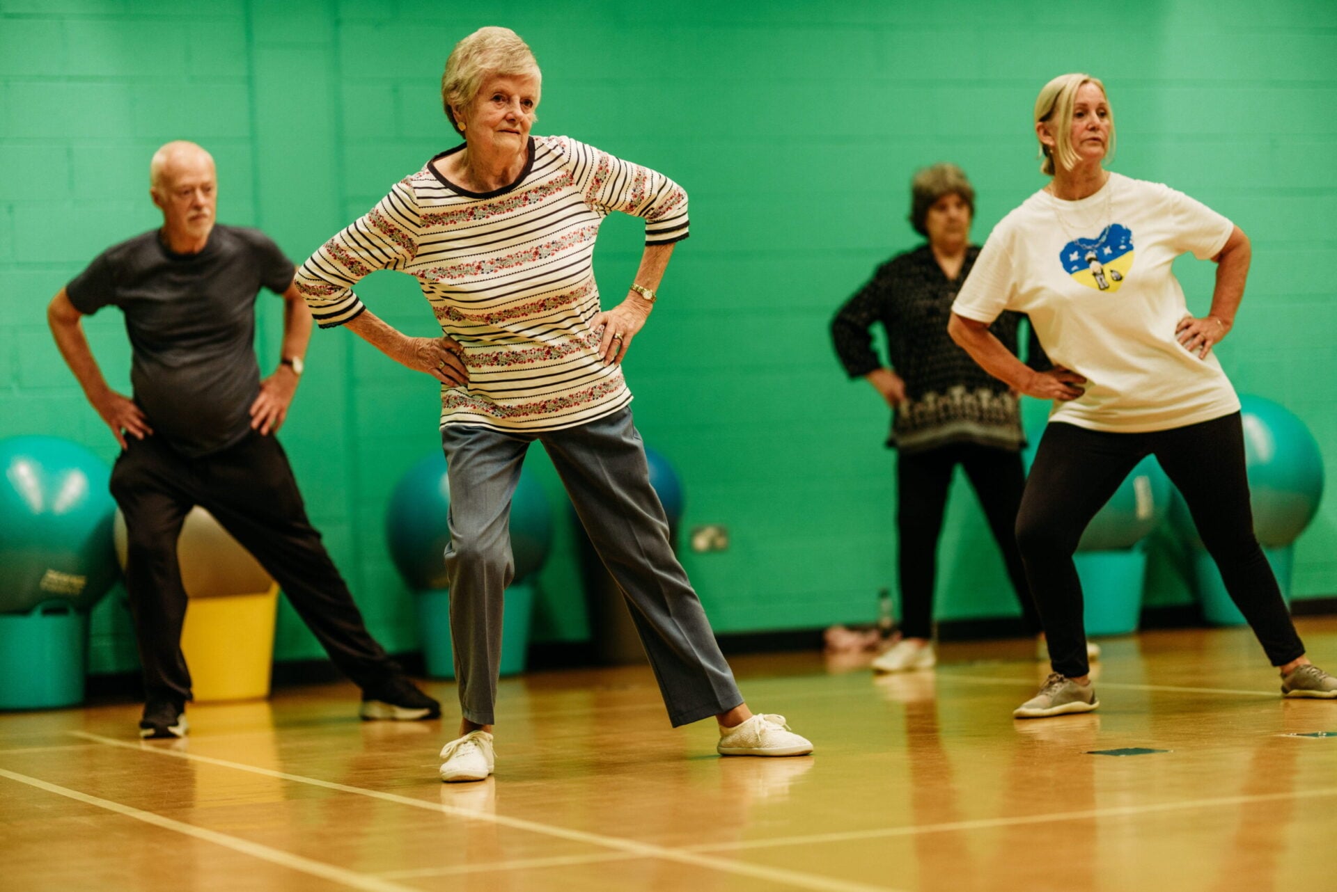 A group of women perform a stretching exercise together during a dance fitness class in a gymnasium.