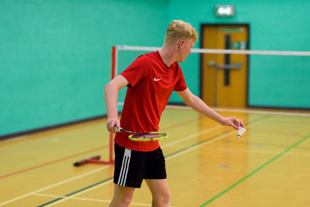 A man in a red shirt plays badminton, reaching to hit the shuttlecock on an indoor court