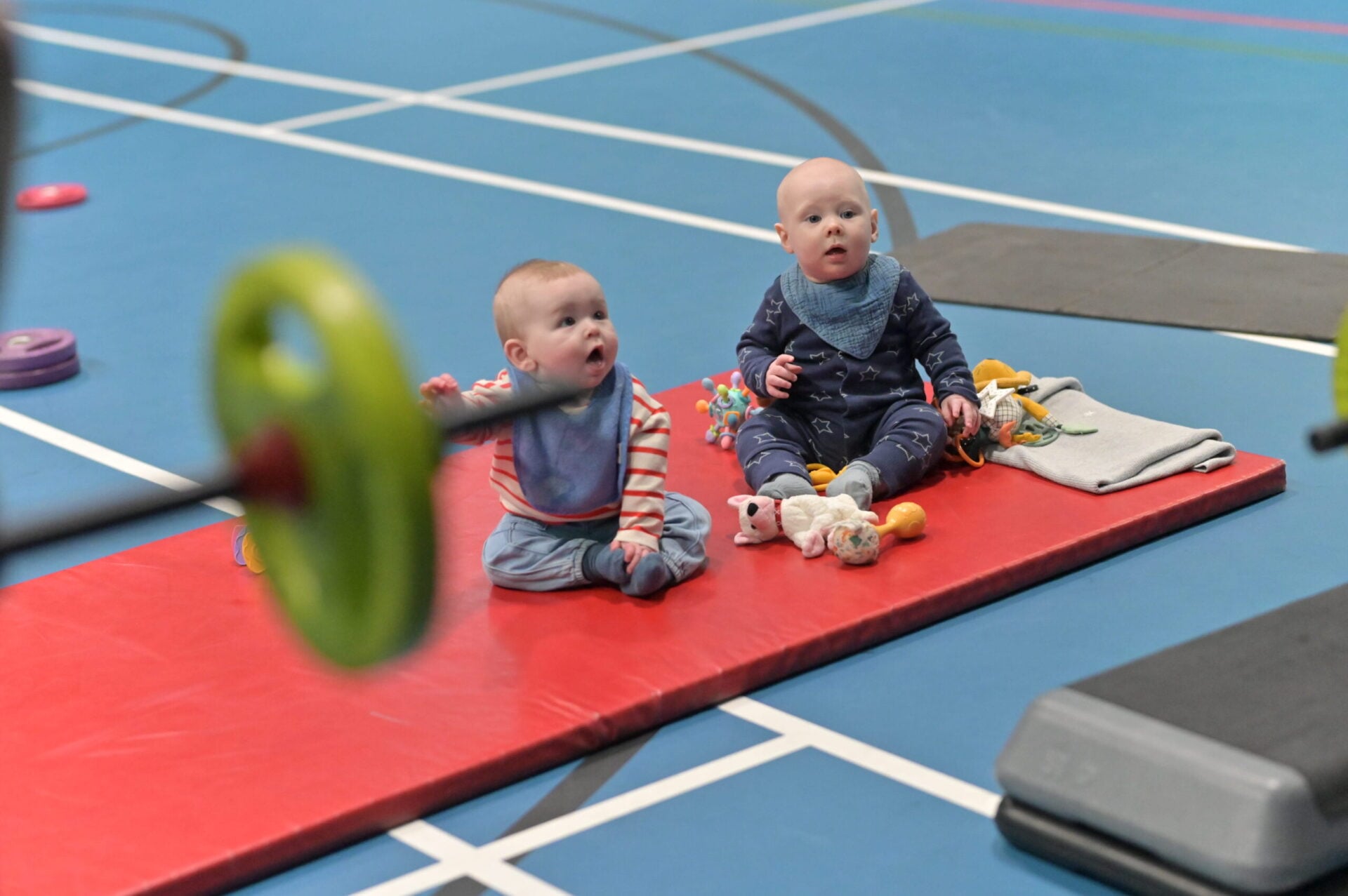 Two babies sit together on a padded mat during a baby gym session.