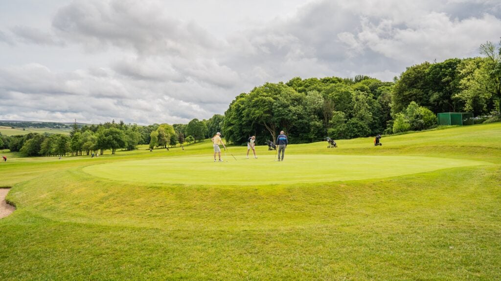 Here are 6 alt text options for this image: "Three golfers putt on a green surrounded by lush trees under a cloudy sky, with golf bags and trolleys nearby