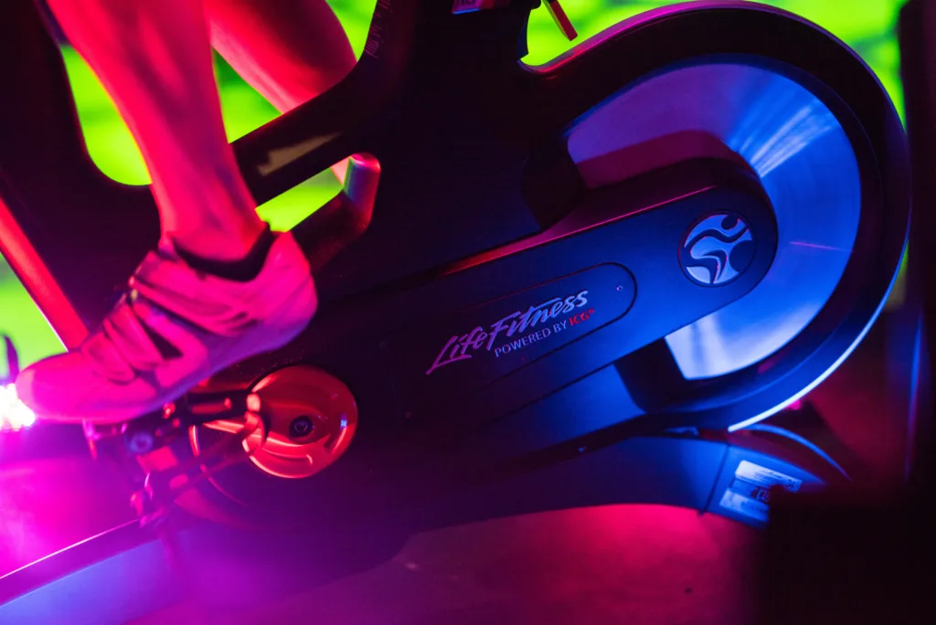 A close-up of a person's foot in athletic shoes clipped into the pedal of a Life Fitness indoor cycling bike, dramatically lit with red, pink, and green lighting, highlighting the bike's flywheel and frame details.
