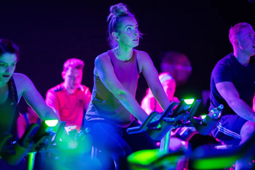 A woman with her hair in a bun rides a stationary bike during an indoor cycling class, illuminated by vibrant blue, purple, and green lighting, with other cyclists visible in the background.